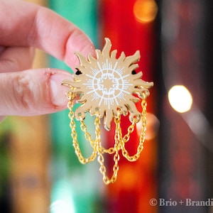 以下が含まれることがあります： 白いディテールとぶら下がっている金色のチェーンが付いた、金色の太陽型のエナメルピン。ピンは中心の星のデザインと太陽光線を特徴としています。ピンは、本とボケライトのぼやけた背景に対して保持されています。