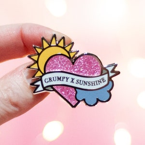 May include: A pink glitter heart with a yellow sun and a blue cloud, with a banner that reads "Grumpy x Sunshine".