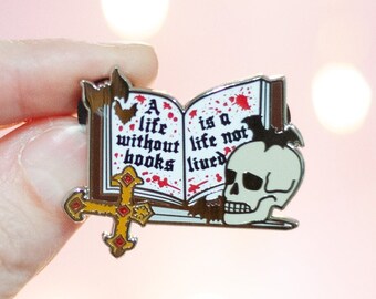 May include: A silver enamel pin featuring an open book with the text "A life without books is a life not lived" and a skull with a cross.