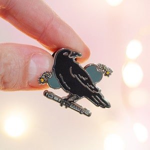 May include: A black enamel pin featuring a crow perched on a silver flute with blue and yellow floral accents.