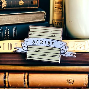 May include: A light blue and brown enamel pin with a scroll design and the word "SCRIBE" in white lettering. The pin is sitting on a stack of books.