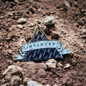 May include: A gold and blue enamel pin with a triangular design and the word "INFANTRY" on a white banner. The pin is on a brown dirt background.
