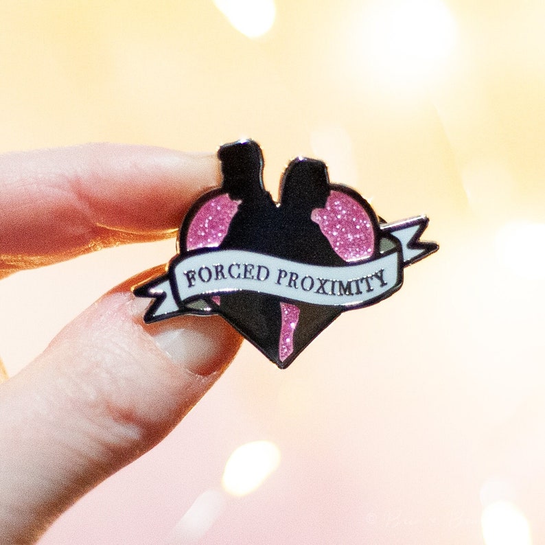 May include: A pink glitter heart-shaped enamel pin with a silhouette of two people dancing inside. The pin has a white banner that says "FORCED PROXIMITY".