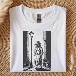 May include: White t-shirt featuring a black and white illustration of a figure in a trench coat and fedora standing in a city alleyway, next to a lamppost. The design has a noir aesthetic.