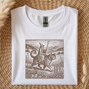 May include: White t-shirt featuring a sepia-toned illustration of a wizard riding a large cat through a field. The wizard wears a pointed hat and holds a staff. The cat has stripes. The shirt is on a woven surface.