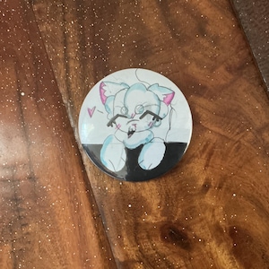 May include: A round button featuring a cartoon cat character with light blue fur, pink ears, and glasses. The cat is drawn with a smiling expression and a small pink heart above its head. The button has a black and white border.