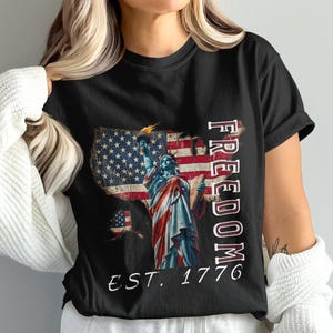 Puede incluir: Camiseta negra con un diseño desgastado de la bandera estadounidense en forma de Estados Unidos y la Estatua de la Libertad. La palabra "FREEDOM" está impresa verticalmente y "EST. 1776" debajo.
