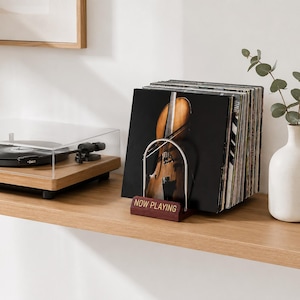 May include: A dark wood vinyl record display stand with a silver metal arch and the text NOW PLAYING in gold lettering. The stand holds a collection of vinyl records, including one with a cello illustration on the cover, next to a turntable.