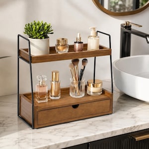 May include: A two-tier wooden bathroom organizer with a black metal frame. The top shelf holds a small potted green plant, a gold jar, a dropper bottle, and a pump bottle. The bottom shelf features a drawer, two perfume bottles, and a glass cup with makeup brushes.