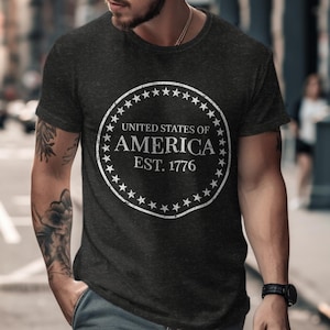 May include: Dark grey t-shirt with a white circular design. The design includes the text "UNITED STATES OF AMERICA EST. 1776" and stars. The shirt is short-sleeved and has a crew neck.