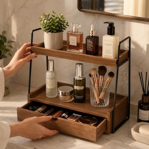 May include: A two-tier wooden bathroom organizer with a black metal frame, featuring a pull-out drawer at the base. The shelves hold various skincare bottles, a small potted plant, a glass cup with makeup brushes, and assorted cosmetics.