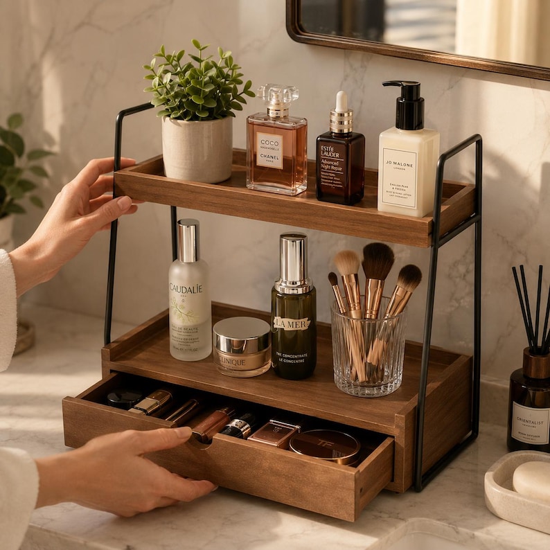 May include: A two-tier wooden bathroom organizer with a black metal frame, featuring a pull-out drawer at the base. The shelves hold various skincare bottles, a small potted plant, a glass cup with makeup brushes, and assorted cosmetics.