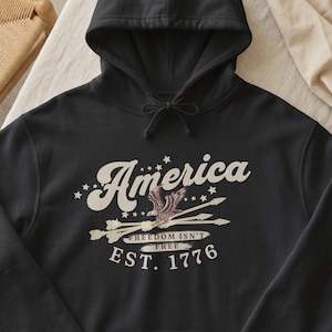 May include: Black hooded sweatshirt with the word "America" in a vintage font, an eagle graphic, and the text "Freedom Isn't Free" and "EST. 1776". The design is in cream and brown tones.