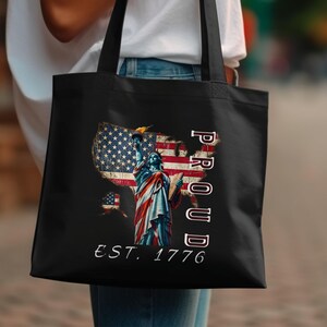 Puede incluir: Bolso tote negro con un gráfico de la Estatua de la Libertad, una bandera estadounidense y la palabra "PROUD" en blanco. También se muestra el texto "EST. 1776". El bolso lo lleva una persona.