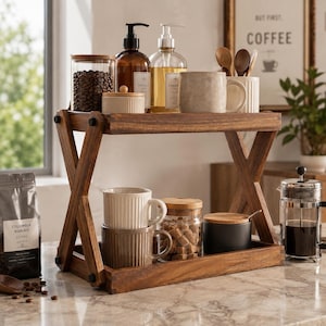 May include: A two-tiered wooden display stand with an X-shaped frame. The top shelf holds a glass jar of coffee beans, two pump bottles, a small lidded container, and a ceramic mug. The bottom shelf holds two stacked mugs, a glass jar of sugar cubes, and a black lidded canister.