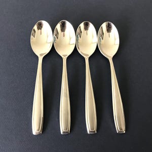 May include: A set of four polished silver-tone metal spoons arranged in a vertical row. Each spoon features a smooth, oval-shaped bowl and a tapered handle with a subtle, raised vertical line detail running down the center of the stem.