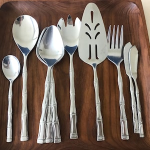 May include: A set of stainless steel flatware featuring handles molded to resemble bamboo stalks. The collection includes a small spoon, a large spoon, two medium spoons, a three-pronged fork, a cake server with cutouts, a dinner fork, and two small knives.