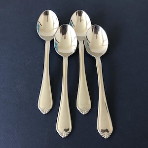 May include: A set of four polished silver-tone metal spoons arranged in a vertical formation. Each spoon features a smooth, rounded bowl and a slender handle that terminates in a decorative, fan-shaped embossed detail at the base of the stem.