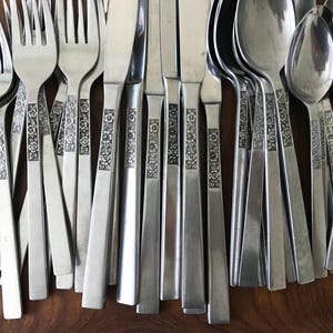 May include: A collection of stainless steel flatware including forks, knives, and spoons. Each piece features a flat handle with a vertical embossed floral pattern running down the center. The utensils are arranged side by side in a uniform row.