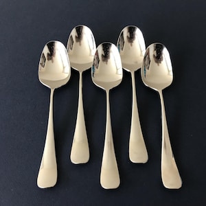 May include: A set of five polished silver-tone metal spoons arranged in a vertical row. Each spoon features a smooth, rounded bowl and a plain, slightly flared handle. The reflective surface of the metal shows light highlights.