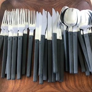 May include: A collection of stainless steel flatware featuring forks, knives, and spoons with matte black plastic handles. The set is arranged in a wooden tray, showcasing the metallic finish of the utensils contrasted against the dark handles.