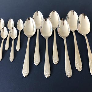 May include: A collection of eleven silver-toned metal spoons arranged in two rows. The spoons feature a polished finish and a decorative fan-like pattern at the base of each handle. The set includes five smaller spoons and six larger spoons.