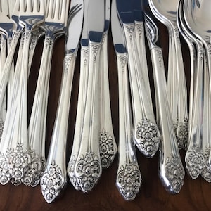 May include: A collection of silver-toned flatware including forks, knives, and spoons. Each piece features a polished finish and an ornate floral embossed pattern at the base of the handle, with vertical fluting extending up the stem.