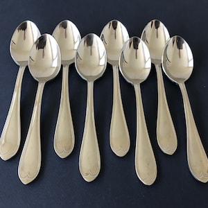 May include: A set of eight polished silver-tone metal spoons arranged in two rows. Each spoon features a smooth, rounded bowl and a tapered handle that ends in a subtle, heart-shaped decorative indentation at the base of the handle.