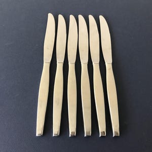 May include: A set of six silver-toned stainless steel dinner knives arranged in a vertical row. Each knife features a smooth, polished finish with a tapered handle and a curved blade edge, presenting a uniform and sleek design for table settings.
