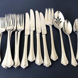 May include: A set of silver-toned stainless steel flatware including four forks, four knives, one large serving fork, one slotted serving spoon, one teaspoon, and two small spoons. Each piece features a smooth handle with a flared base.