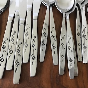 May include: A collection of stainless steel flatware including knives, forks, and spoons. Each piece features a slender handle decorated with a repeating etched floral pattern consisting of small four-petaled flowers and thin stems.