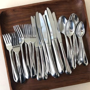 May include: A collection of stainless steel flatware including forks, knives, and spoons arranged on a square wooden tray. The set features polished silver-toned metal utensils with smooth, rounded handles and classic tines on the forks.