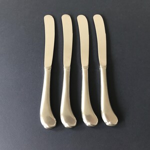 May include: A set of four silver-toned butter knives arranged vertically. Each knife features a smooth, polished finish with a rounded, slightly curved handle and a blunt, rounded blade tip, reflecting a clean and uniform metallic appearance.