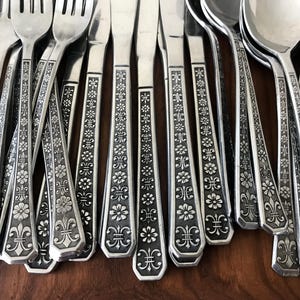May include: A collection of stainless steel flatware including forks, knives, and spoons. Each piece features a detailed embossed pattern on the handle consisting of repeating floral motifs and fleur-de-lis accents with a darkened finish.