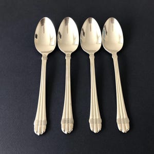 May include: Four silver-toned metal spoons arranged in a row. Each spoon features an oval bowl and a handle decorated with three vertical ridges and a horizontal banded detail near the base, which ends in a rounded, scalloped edge design.