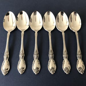 May include: A set of six silver-toned metal spoons arranged in a row. Each spoon features a polished oval bowl and a decorative handle with intricate embossed floral and scrollwork detailing extending from the neck down to the rounded base.