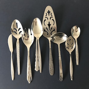 May include: A collection of eight silver-toned metal serving utensils including a cake server, a ladle, slotted spoons, and various forks and knives. Each piece features distinct decorative patterns on the handles and functional heads.