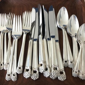 May include: A collection of polished silver-plated flatware including forks, knives, and spoons. Each piece features a decorative floral motif at the base of the handle. The set is arranged together, showcasing the reflective metallic finish.