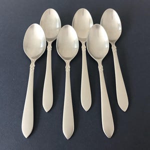 May include: A set of six silver-toned metal spoons arranged in two rows. Each spoon features a smooth, rounded bowl and a tapered handle with a subtle, decorative textured detail at the neck where the handle meets the bowl of the spoon.