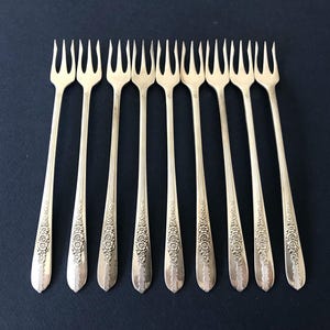May include: A set of nine silver-toned metal cocktail forks arranged in a parallel row. Each fork features a slender handle with an embossed floral pattern at the base and three long, thin tines at the top, designed for serving small appetizers.
