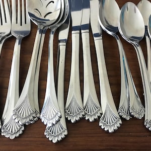 May include: A collection of stainless steel flatware including forks, knives, and spoons. Each piece features a polished finish and a decorative fan-shaped motif at the base of the handle. The set includes a slotted serving spoon among the utensils.