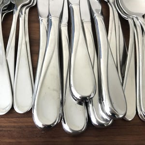 May include: A collection of polished stainless steel flatware pieces, including forks, knives, and spoons, arranged in a cluster. Each utensil features a smooth, rounded handle with a subtle, curved indentation running along the length of the metal.