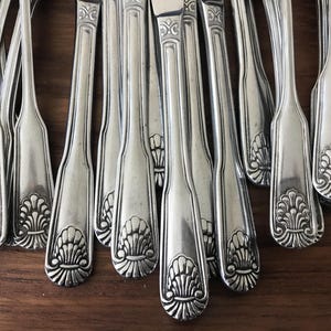 May include: A collection of silver-toned metal flatware handles arranged in a vertical row. Each handle features a polished finish and a decorative embossed shell motif at the base, with a slender neck and a subtle raised border along the edges.