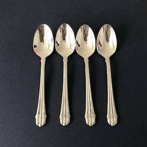 May include: A set of four polished silver-tone metal spoons arranged in a row. Each spoon features an oval bowl and a slender handle decorated with vertical fluted lines and a horizontal banded detail near the base of the handle.