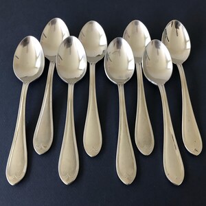 May include: A set of eight polished silver-tone metal spoons arranged in two rows. Each spoon features a smooth, oval-shaped bowl and a slender handle that widens toward the base, ending in a subtle, rounded heart-shaped decorative detail.