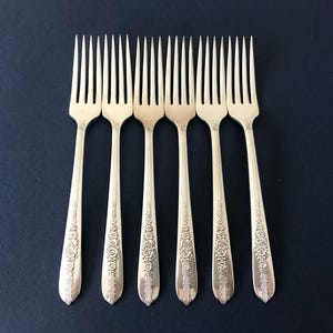 May include: A set of six silver-toned metal forks arranged in a row. Each fork features a slender handle with an embossed floral pattern near the base and four long, thin tines. The metal has a polished finish with reflective surfaces.