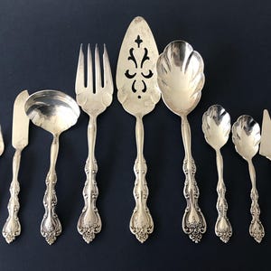 May include: A set of eight silver-toned serving utensils featuring ornate, embossed handles with floral detailing. The collection includes a small fork, a butter knife, a ladle, a large serving fork, a cake server, a large spoon, two smaller spoons, and a triangular spreader.
