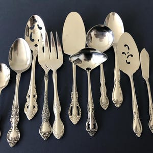 May include: A collection of ten silver-toned metal serving utensils and spoons featuring ornate, embossed floral handles. The set includes a small spoon, two large spoons, a slotted spoon, a two-pronged fork, a cake server, two ladles, a slotted spatula, and a butter knife.
