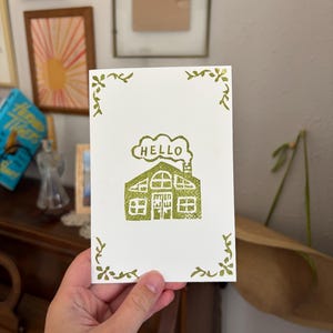 May include: A white greeting card with a green glittery illustration of a house and the word "HELLO". The card has a decorative green border. The card is held in a person's hand.