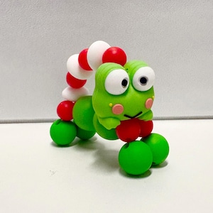May include: A green cartoon frog figurine with large white eyes, pink cheeks, and a red mouth. The frog has a segmented body with red, white, and green spheres. The figurine is on a white surface.